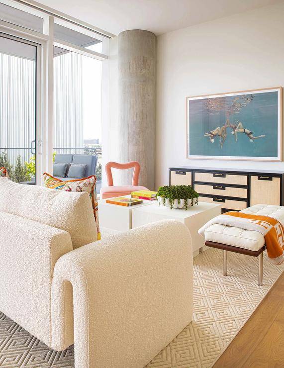 An ivory boucle sofa placed on a beige diamond pattern rug faces styled white pedestal bunched coffee tables matched with a mid-century modern leather bench draped in an orange Hermes throw blanket.