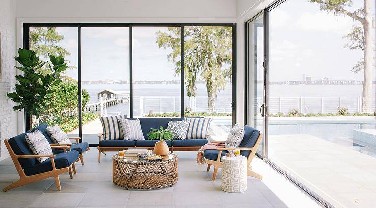 Brown and blue mid century modern lounge chairs on gray tiles join a round wood and rope metal coffee table in a sunroom with glass doors and windows.